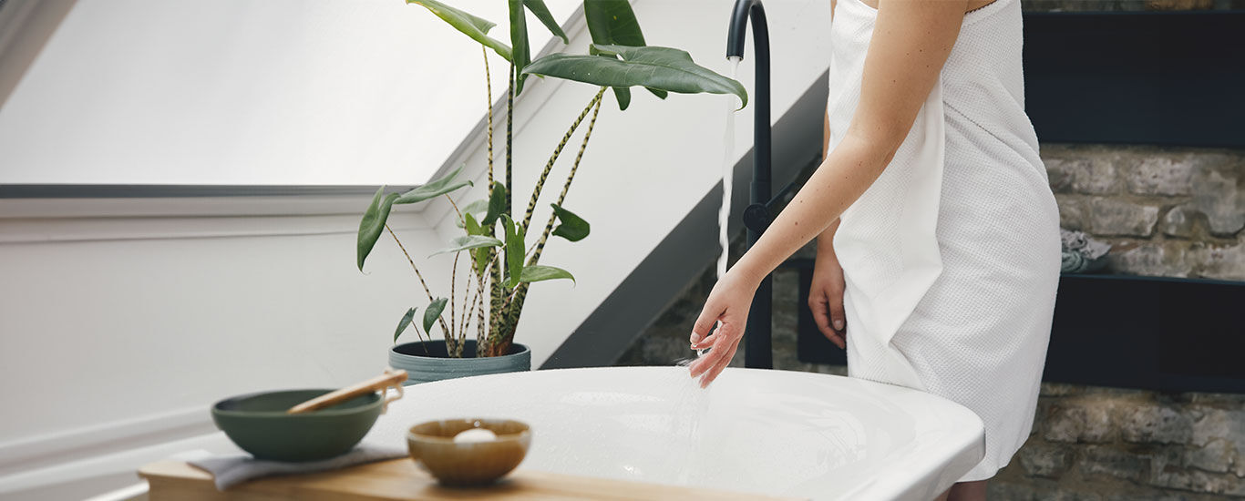 Frau im Handtuch steht neben der Badewanne und überprüft Badewassertemperatur. 