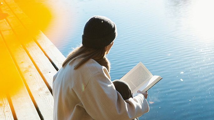 Frau sitzt in der Sonne am Seesteg und ließt ein Buch.