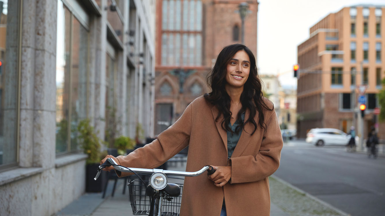 Frau steht mit einem Fahrrad auf einem Gehweg in einer Stadt, umgeben von hohen Gebäuden und einer Straße mit Autos im Hintergrund.