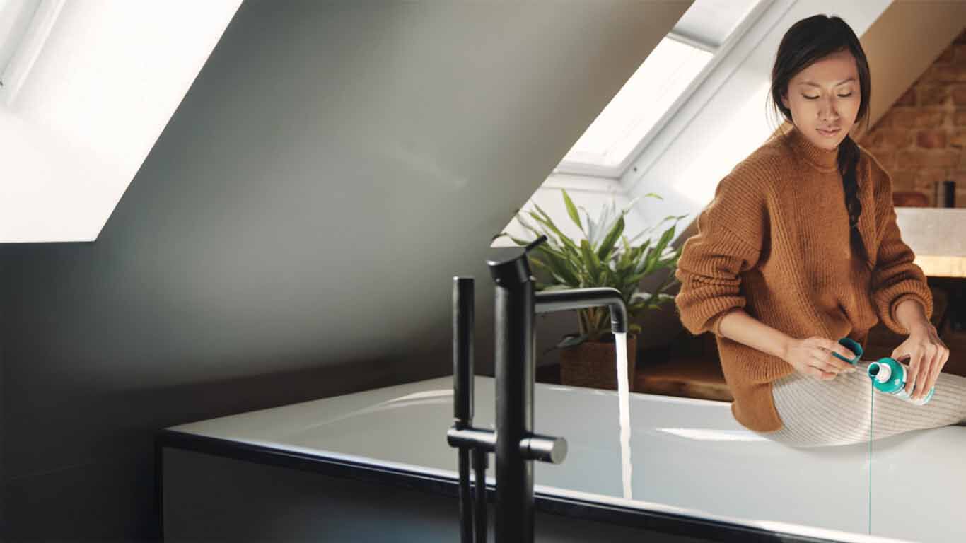  Wasser läuft  in eine Badewanne ein, auf dem Badewannenrand sitzt eine Frau die ein Badeinfüllt.