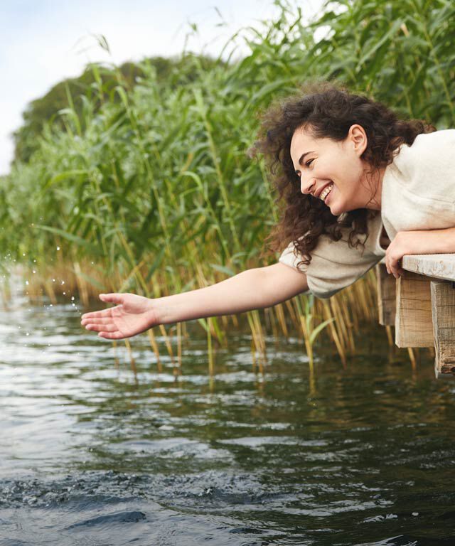 Frau liegt auf einem Steg am Wasser und streckt ihre Hand in Richtung Wasser aus. 
