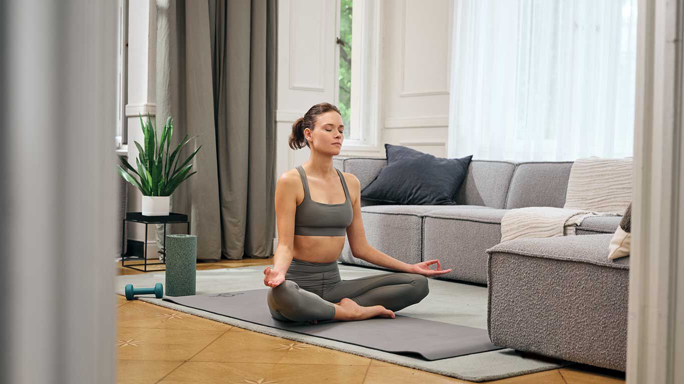 Frau im Schneidersitz auf Yogamatte im Wohnzimmer.