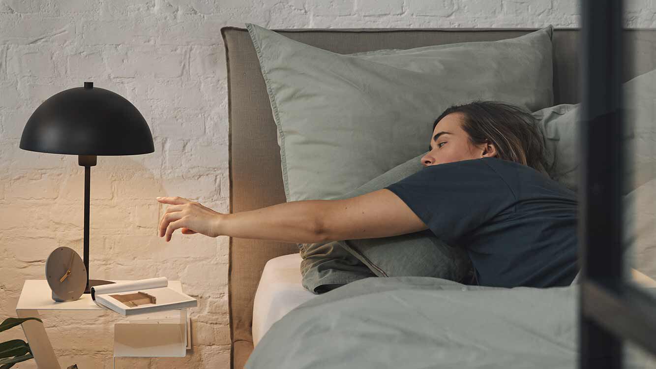 Frau liegt im Bett und streckt sich nach Nachttischlampe.