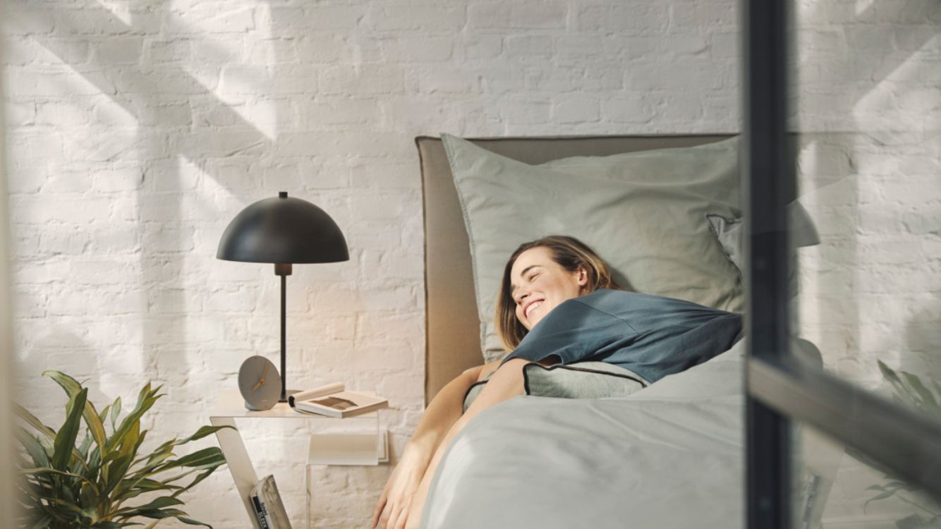  Frau liegt im Bett, l&auml;sst ihre Arme vom Bett h&auml;ngen und schaut in die Sonnenstrahlen.