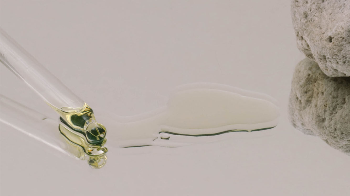 Pipette mit einem Tropfen klarer Flüssigkeit neben einem porösen Stein auf spiegelnder Oberfläche.