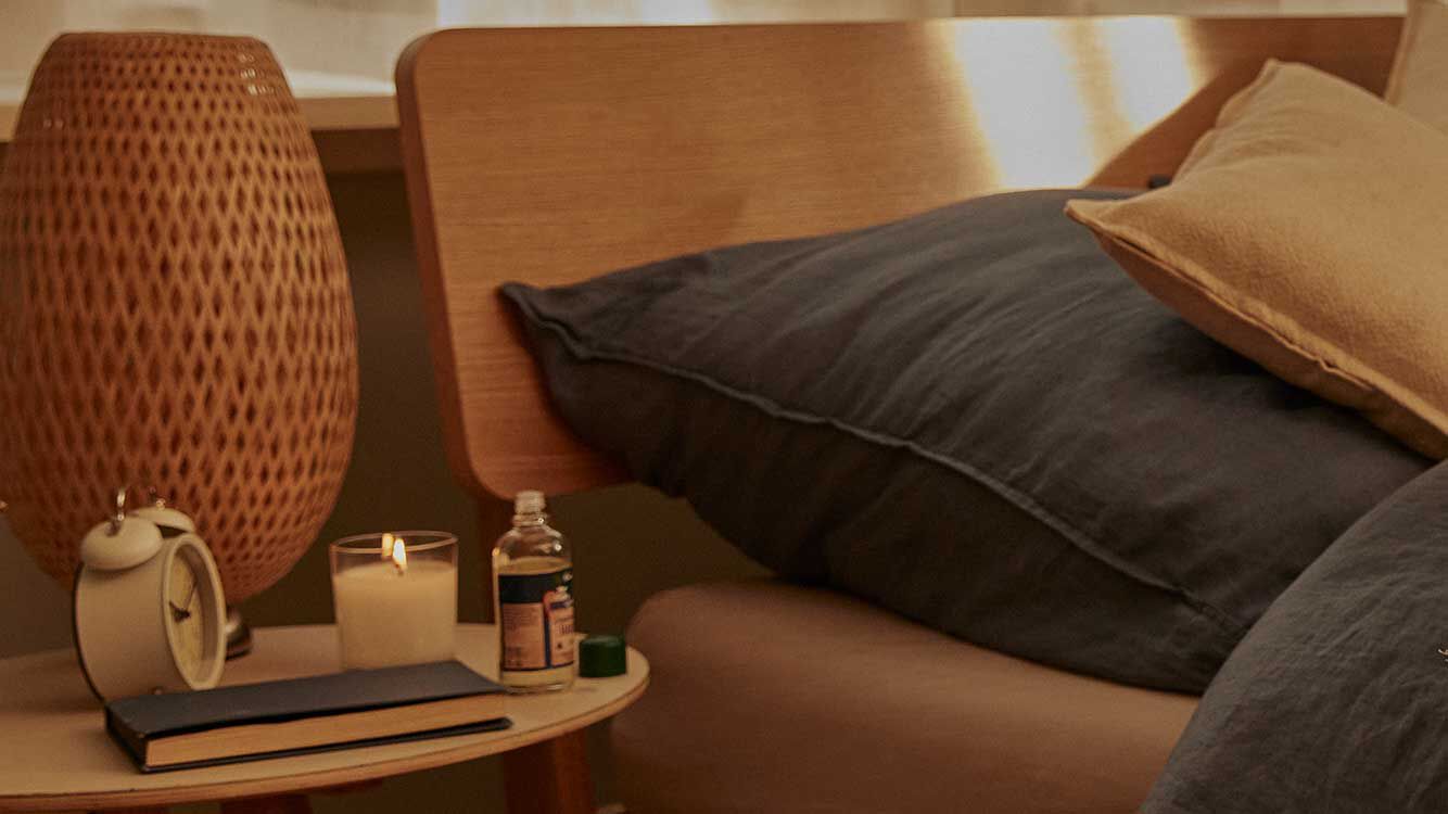 Wecker, Kerze und Kneipp Gute Nacht Öl stehen auf einem Nachttisch. Im Hintergrund ein Bett.