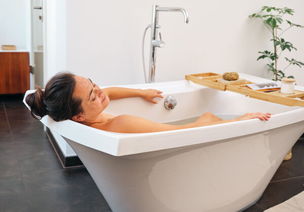 Dunkelhaarige Frau mit Dutt liegt in der Badewanne und entspannt. 