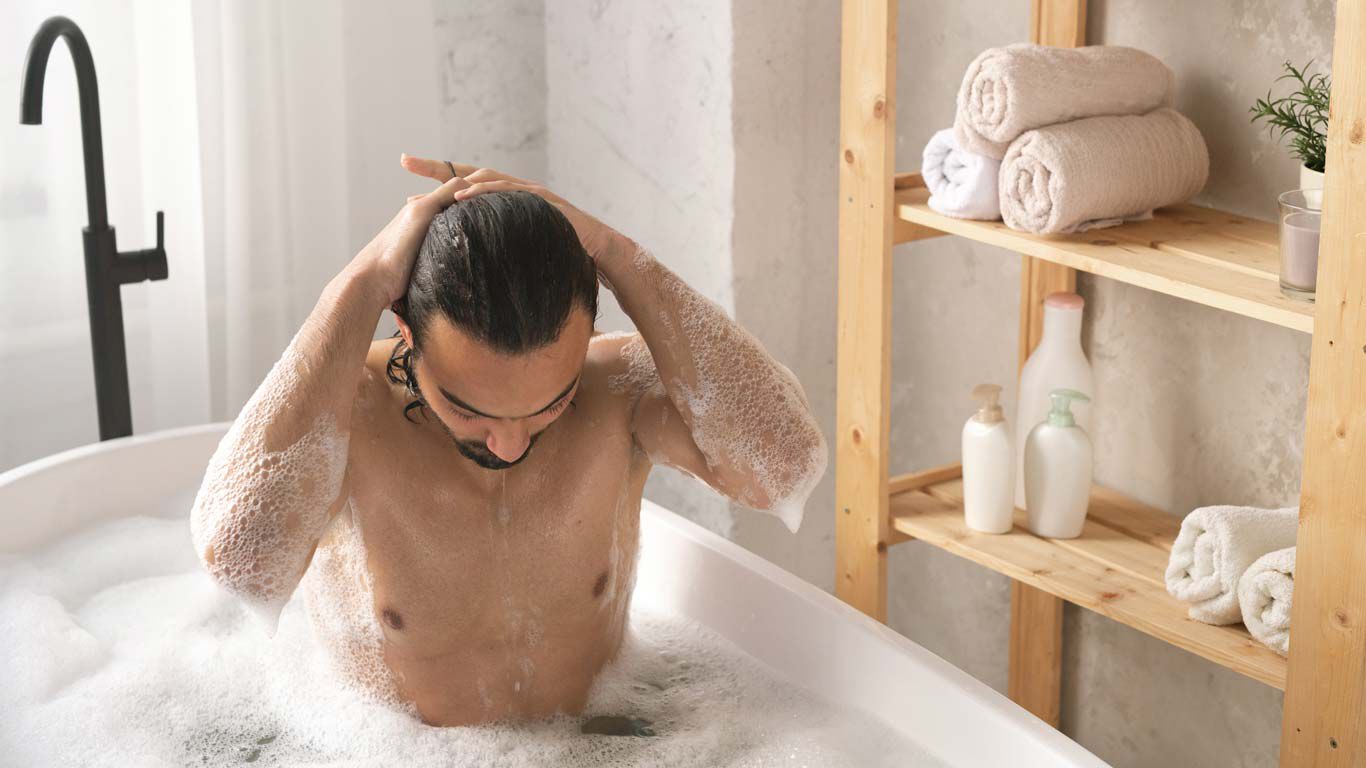 Mann sitzt in der Badewanne und streift sich die Haare zurück.