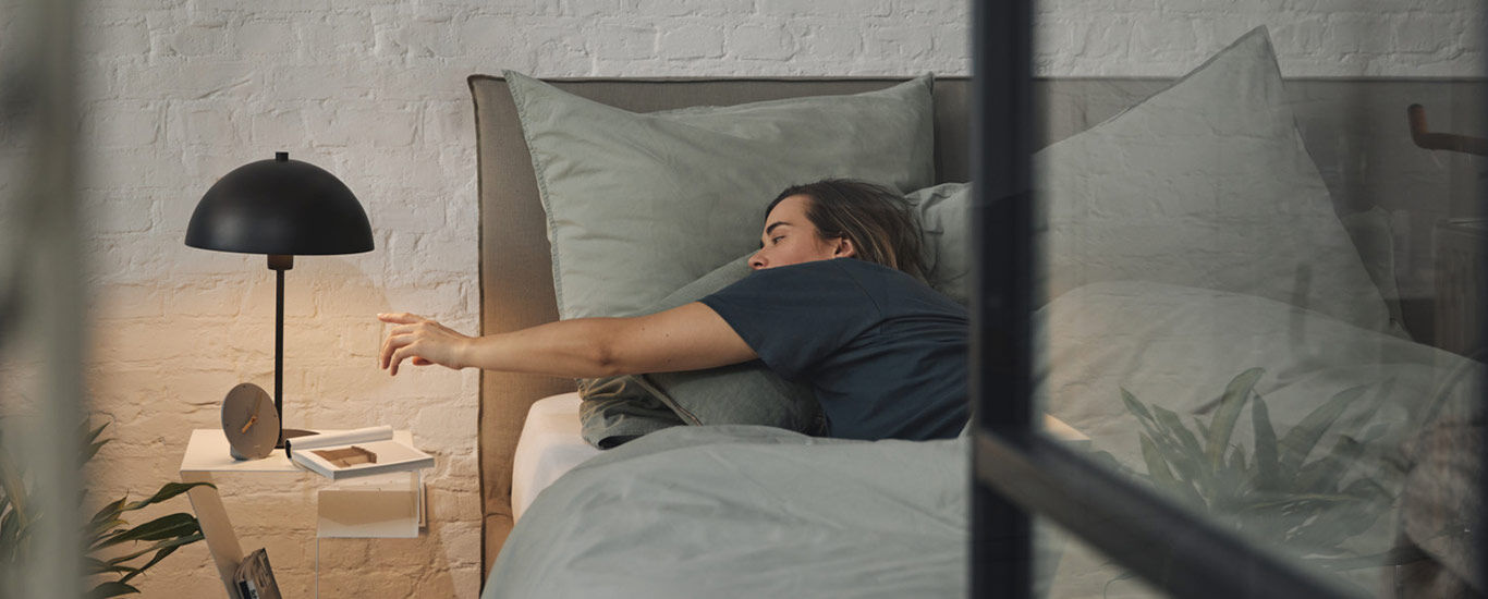 Frau liegt im Bett und streckt sich in Richtung Nachttisch
