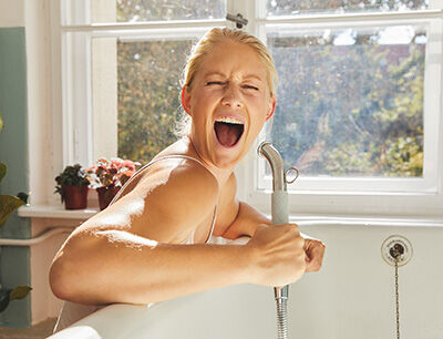 Frau singt an der Badewanne mit der Brause als Mikrophon