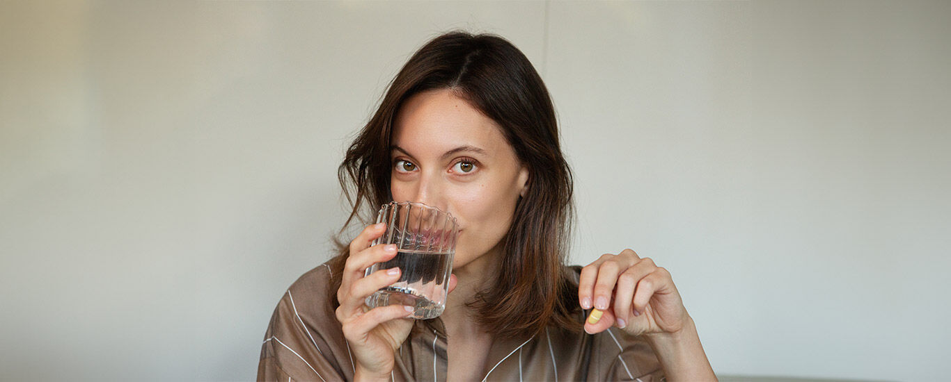 Frau trinkt ein Glas Wasser und hält Nahrungsergänzungsmittel in der Hand. 