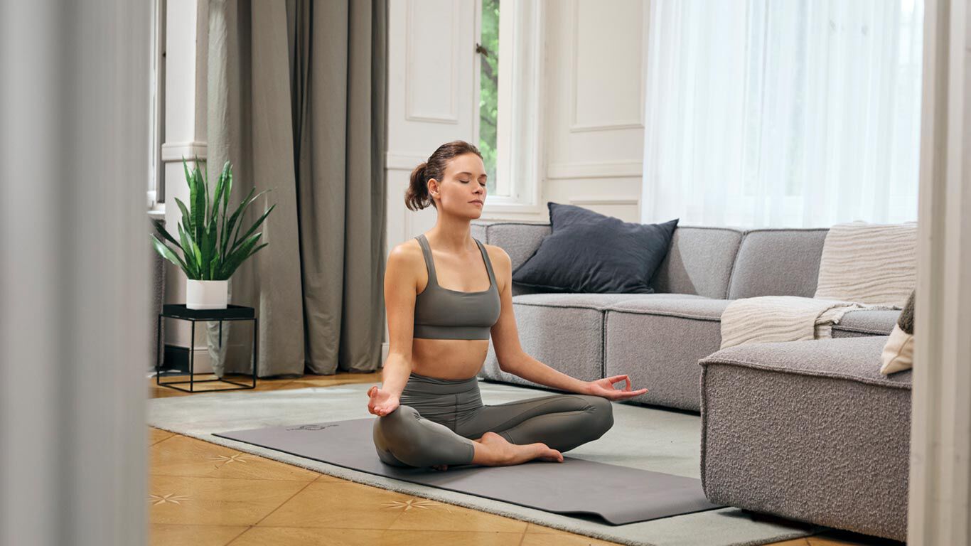 Frau sitzt im Schneidersitz auf einer Yogamatte und meditiert in einem Wohnzimmer vor einem Sofa.