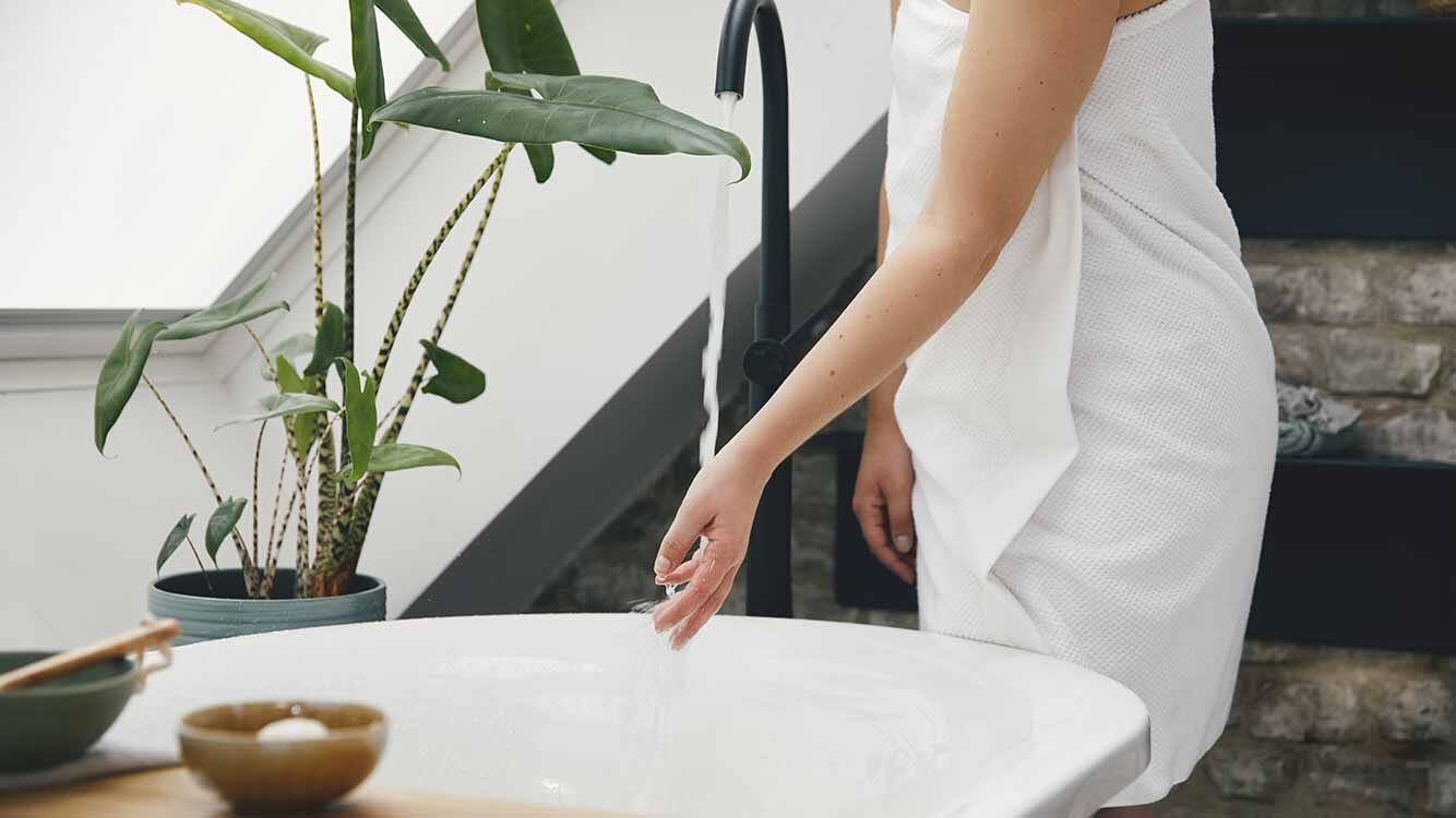 Frau im Handtuch steht neben der Badewanne und überprüft Badewassertemperatur. 