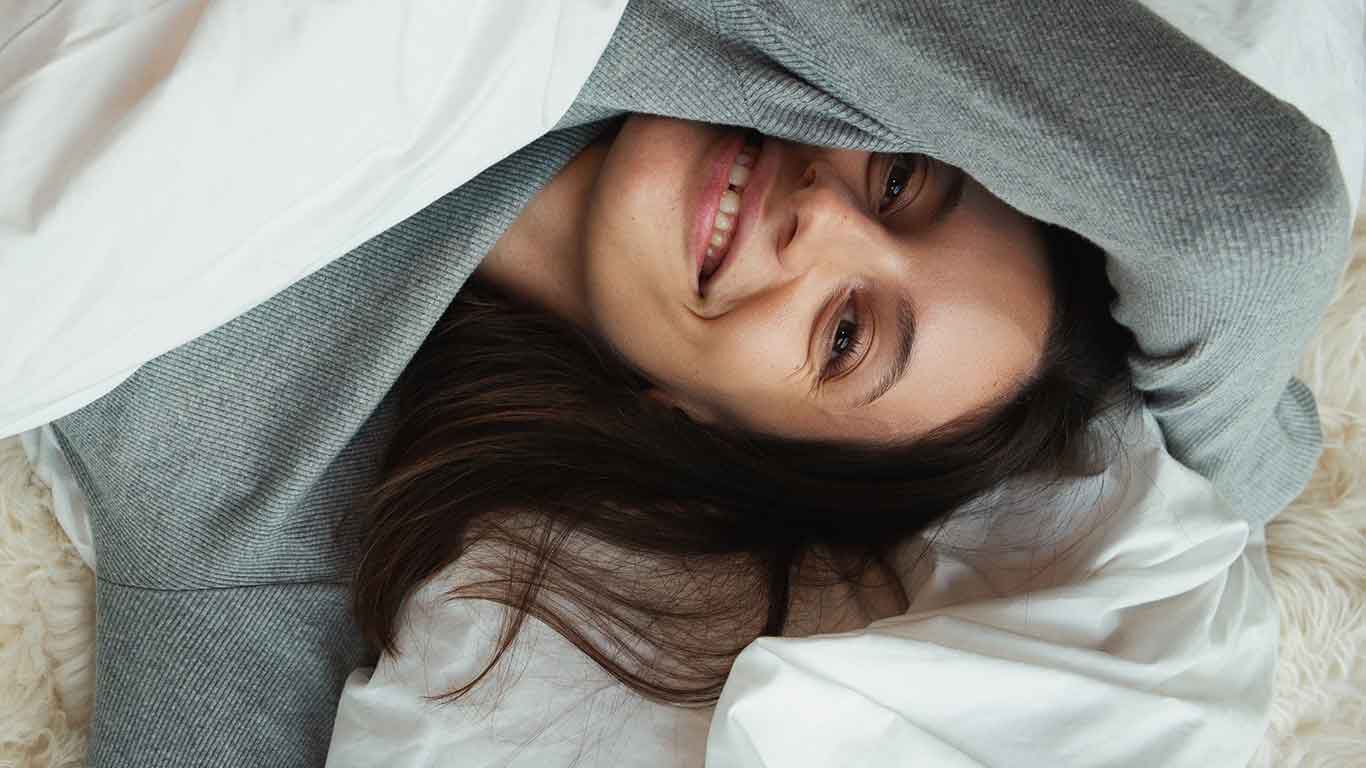Frau liegt eingekuschelt in Bettdecke und lacht in die Kamera.