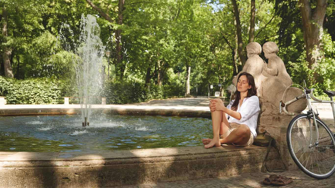 Frau sitzt an einem Brunnen angelehnt und hat die Augen geschlossen, neben ihn ihr Fahrrad anlehnend an den Brunnen