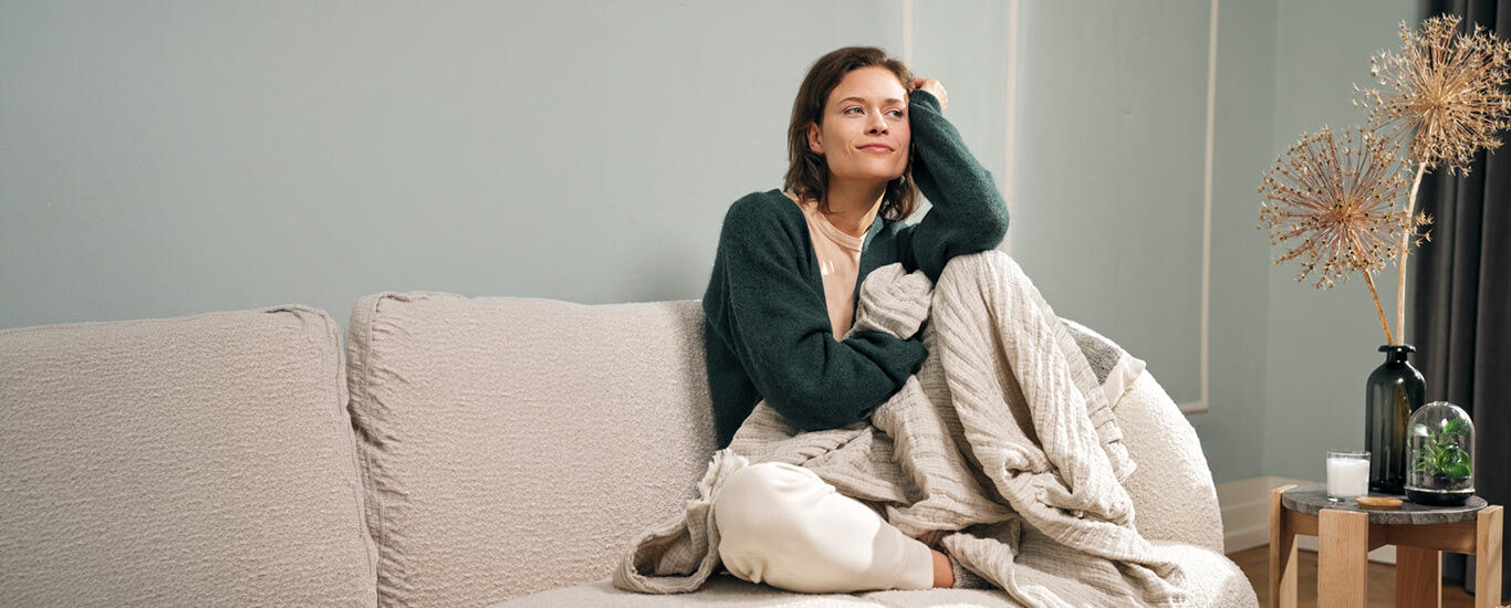 Frau sitzt mit einer Decke auf einem Sofa im Wohnzimmer und st&uuml;tzt sich auf 