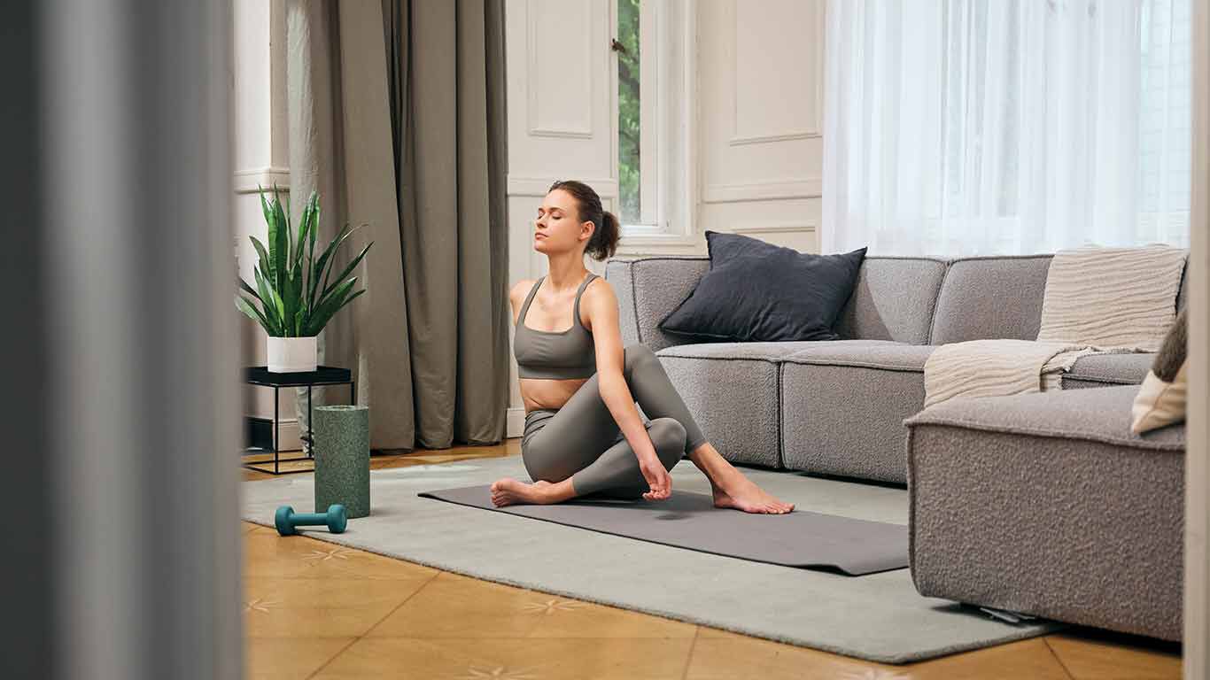 Frau in Yogaklamotten übt drehenden Sitz auf Yogamatte im Wohnzimmer aus.