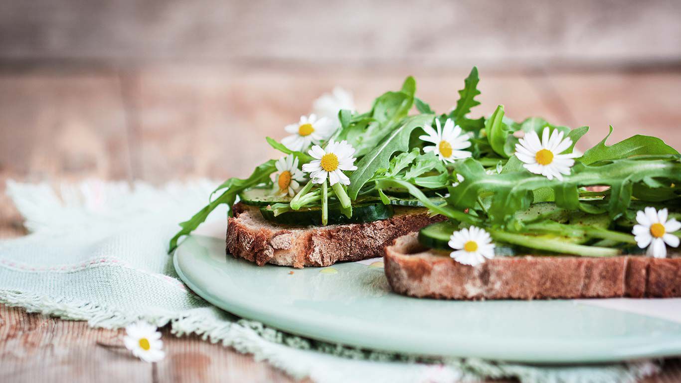 Zwei Scheiben Brot auf einem Teller mit Rucola und Gänseblümchen. 