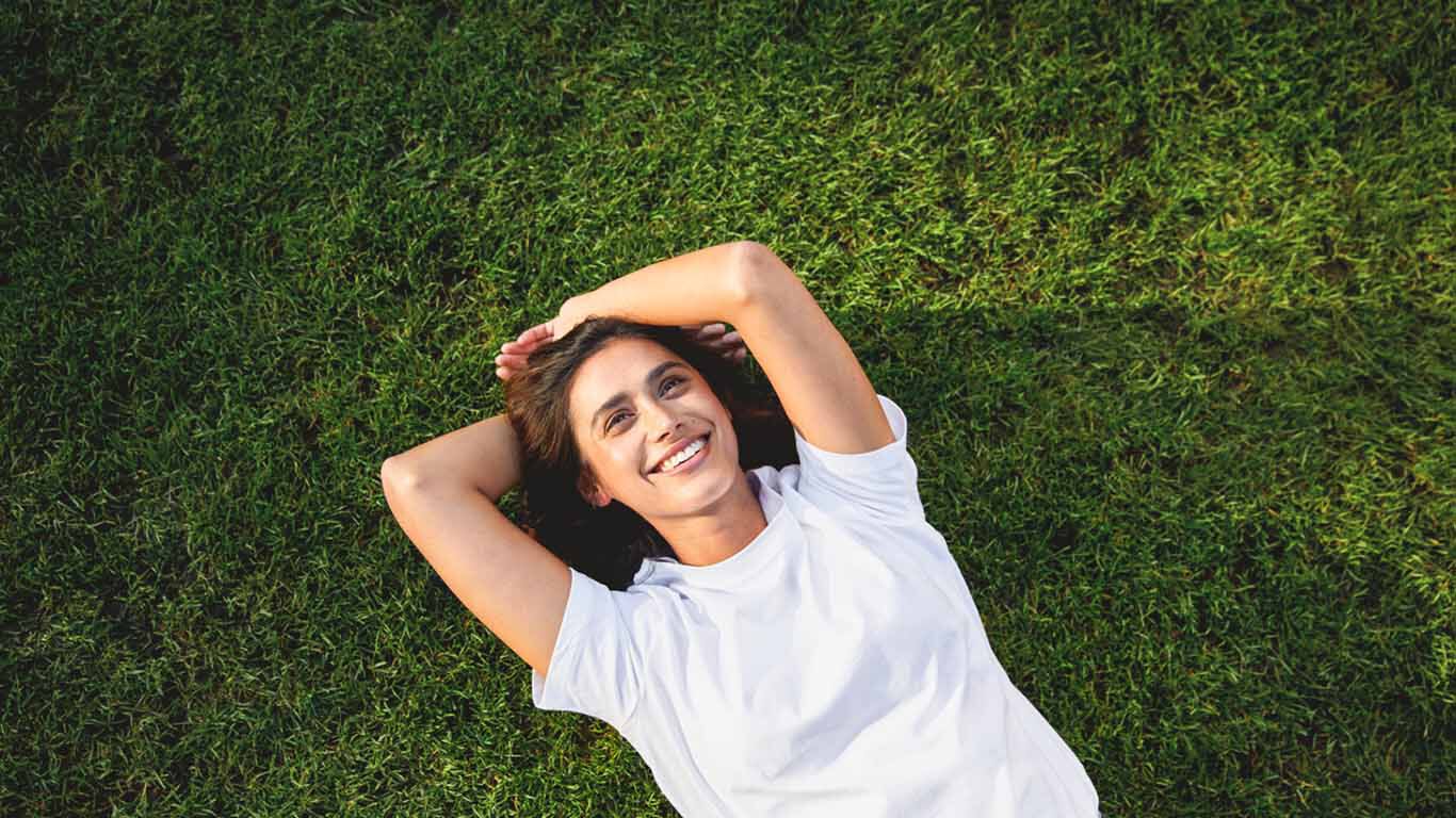 Frau liegt lachend im Gras mit &Uuml;berschlagenen H&auml;nden &uuml;ber dem Kopf und schaut in die Sonne.