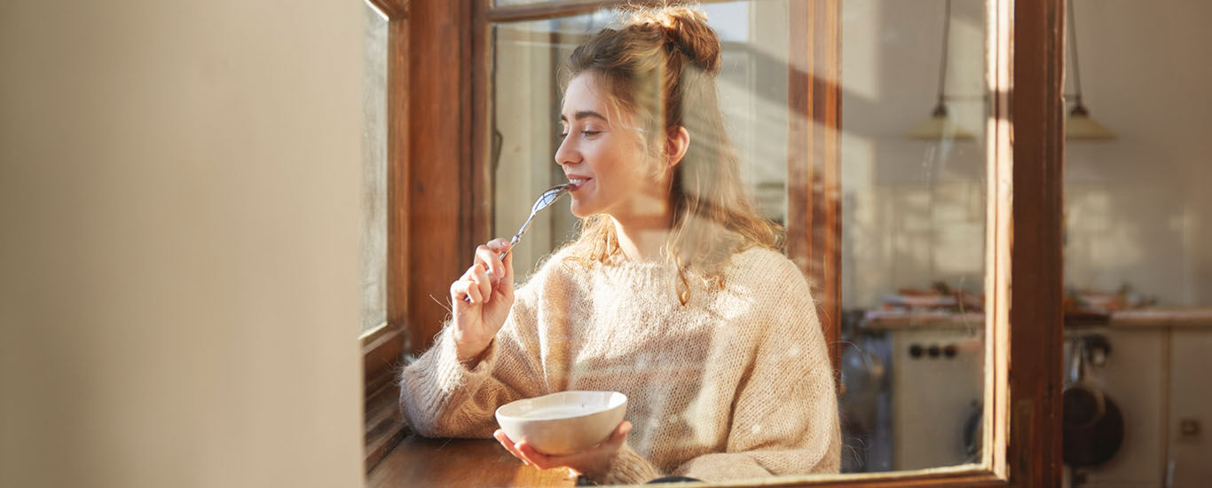 Frau sitzt mit gesunden Fr&uuml;hst&uuml;ck am Fenster mit einer Sch&uuml;ssel M&uuml;sli. Nat&uuml;rliches Tageslicht und eine entspannte Atmosph&auml;re.