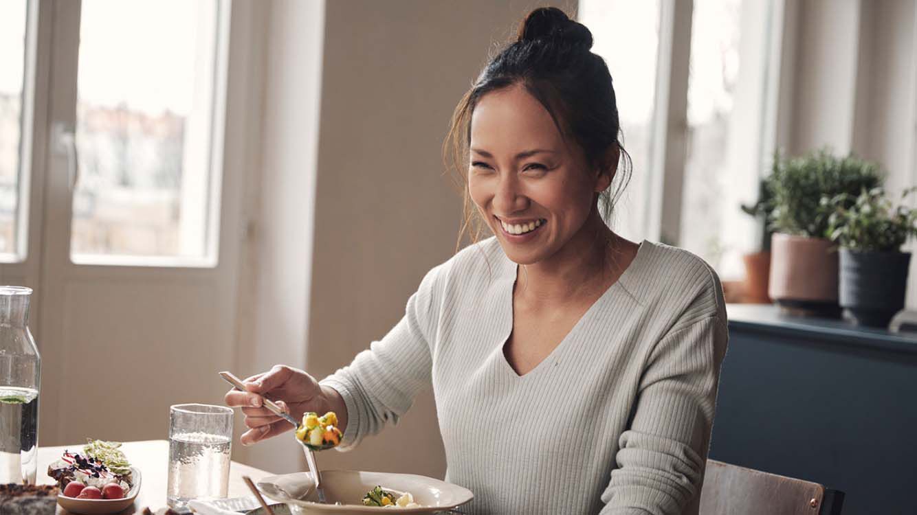 Frau mit Dutt sitzt am Essenstisch, h&auml;lt einen L&ouml;ffel in der Hand und lacht herzlich.