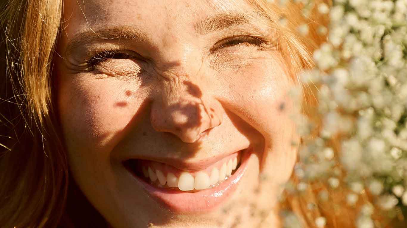 Blonde Frau strahlt lächelnd in die Kamera. Auf ihrem Gesicht spiegeln sich Pflanzenschatten. 
