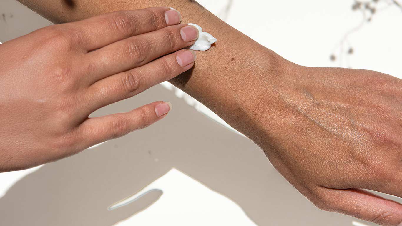 Nahaufnahme von zwei Händen vor hellem Hintergrund. Rechte Hand trägt Creme auf linkes Handgelenk auf.