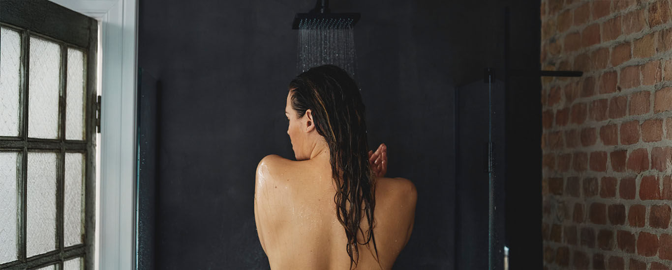 Frau steht unter einer laufenden Dusche in einem modernen Badezimmer, R&uuml;ckenansicht.