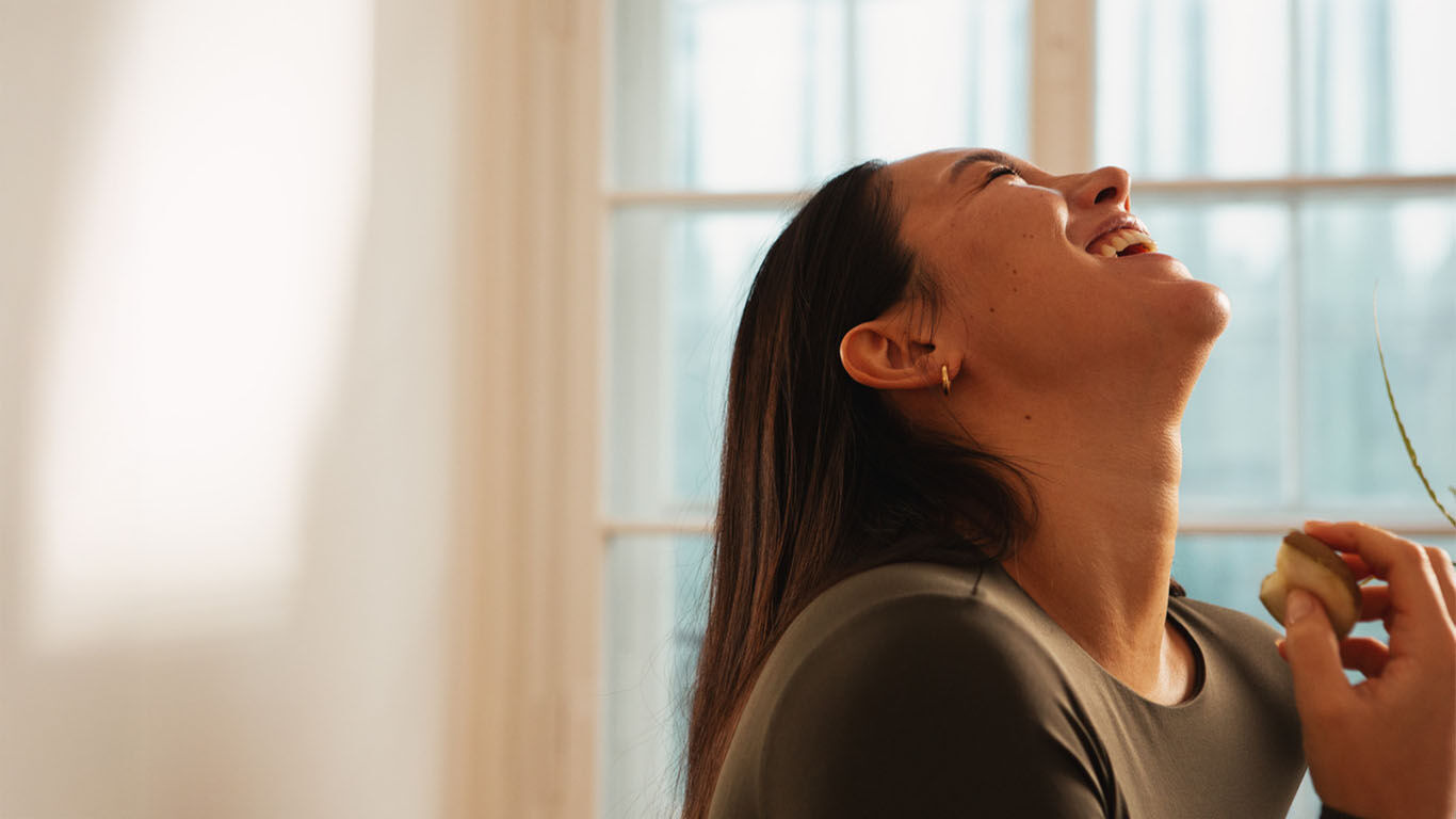 Frau in Jeansjacke steht lachend in der Natur am Strand. 