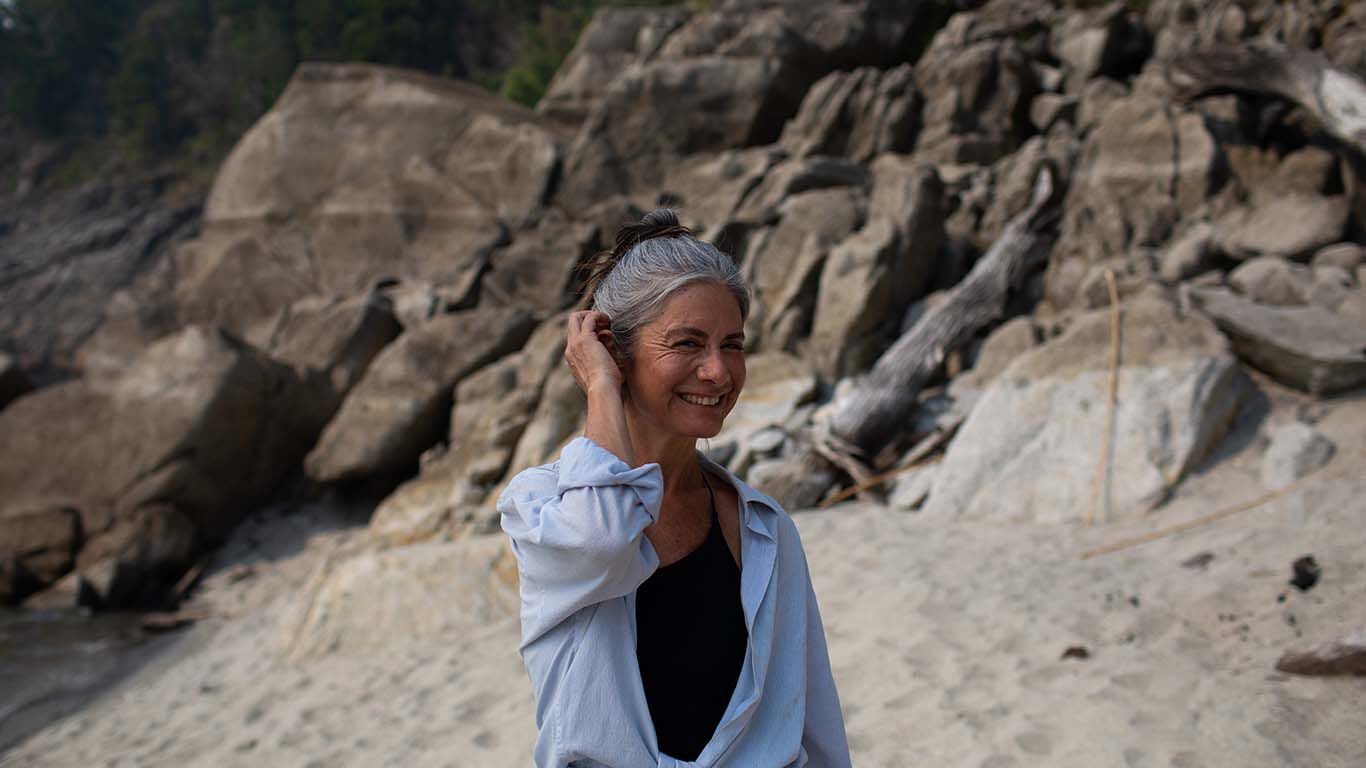 Dame steht am strand vor einer Felswand und legt sich einen Haarsträhne hinter ihr Ohr