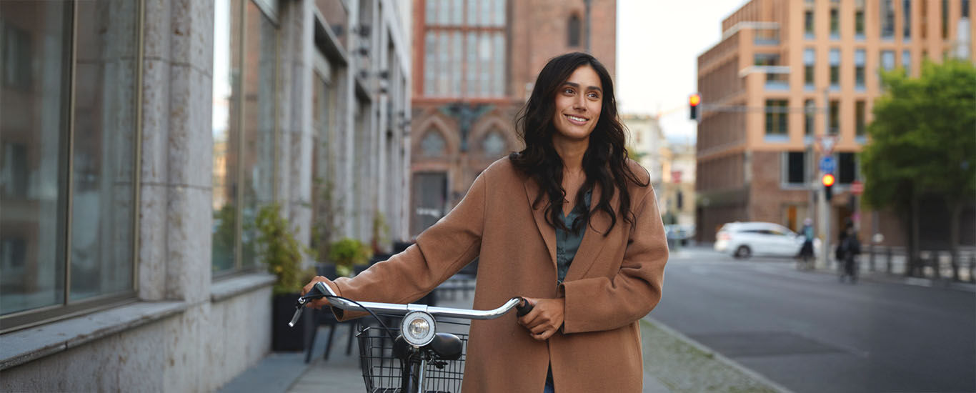 Frau steht mit einem Fahrrad auf einem Gehweg in einer Stadt, umgeben von hohen Gebäuden und einer Straße mit Autos im Hintergrund.
