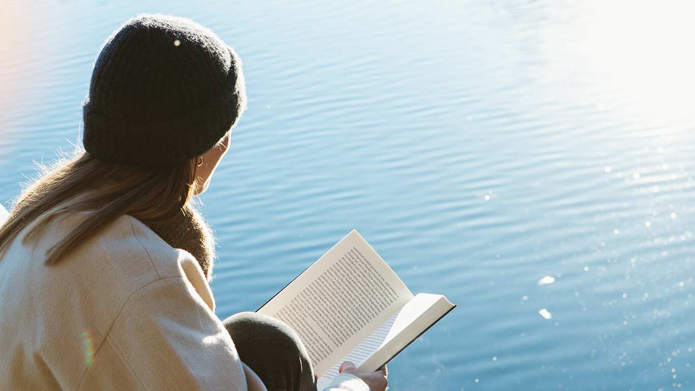 Frau sitzt mit Buch am Wasser in der Sonne. 