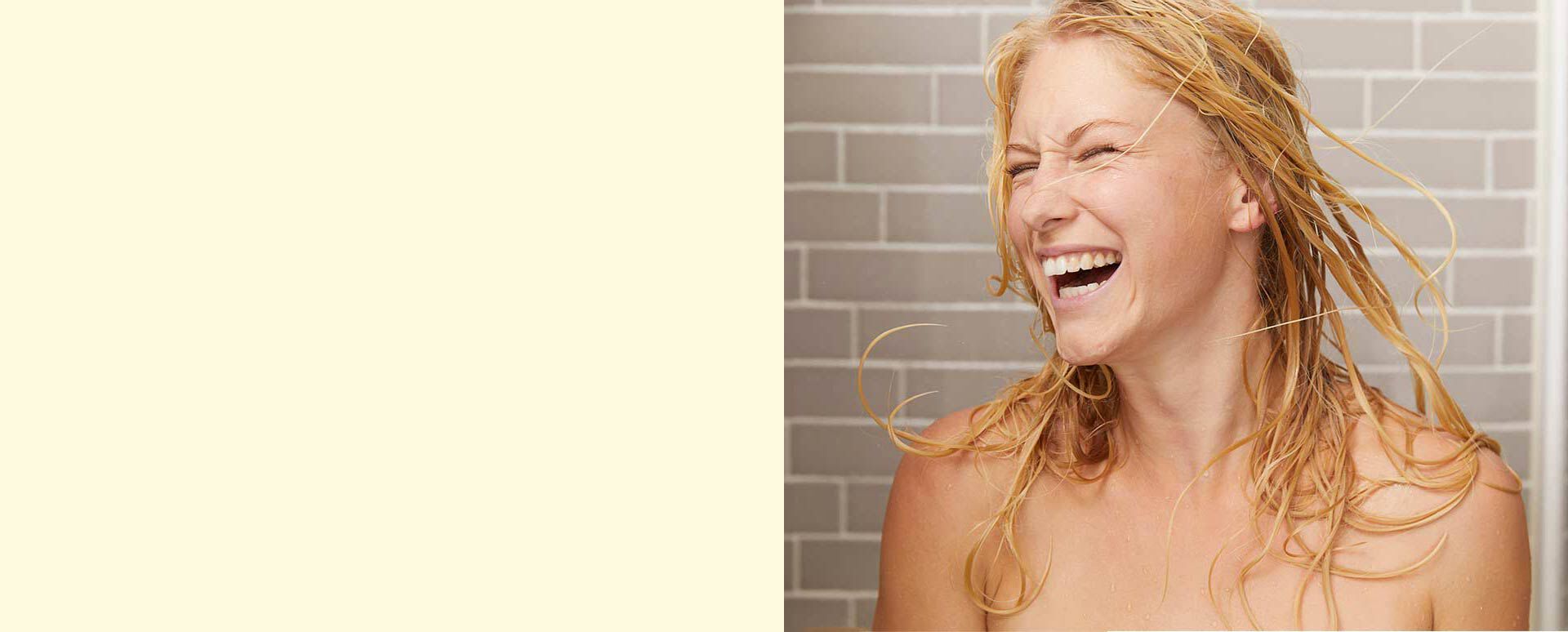 Frau mit blonden nassen Haare steht glücklich unter der Dusche. 