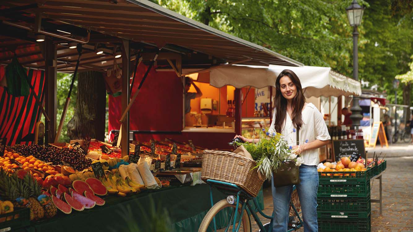 Frau mit Fahrradkorb voller Gem&uuml;se auf einem Wochenmarkt vor Obst- und Gem&uuml;sest&auml;nden.