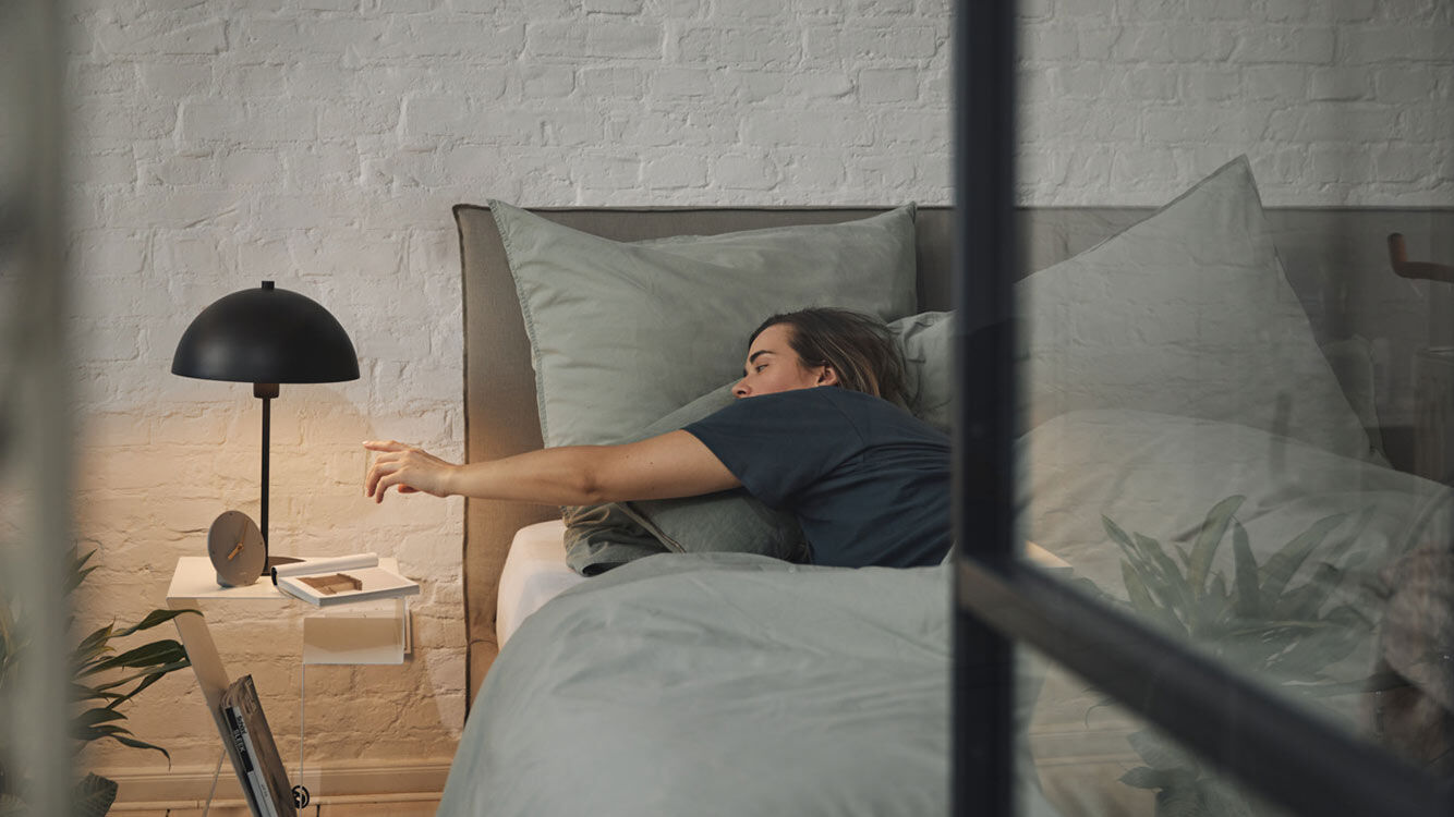 Frau liegt im Bett und streckt sich in Richtung Nachttisch