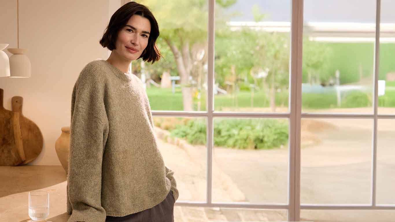 Frau in braunem Strickpullover steht entspannt an der K&uuml;chenzeile vor einer Fensterfront.