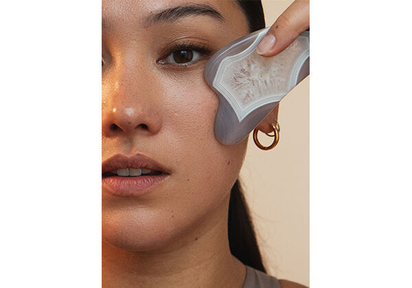 Schwarzhaarige Frau im Top f&uuml;hrt einen Gua Sha Stein &uuml;ber ihr Gesicht. 