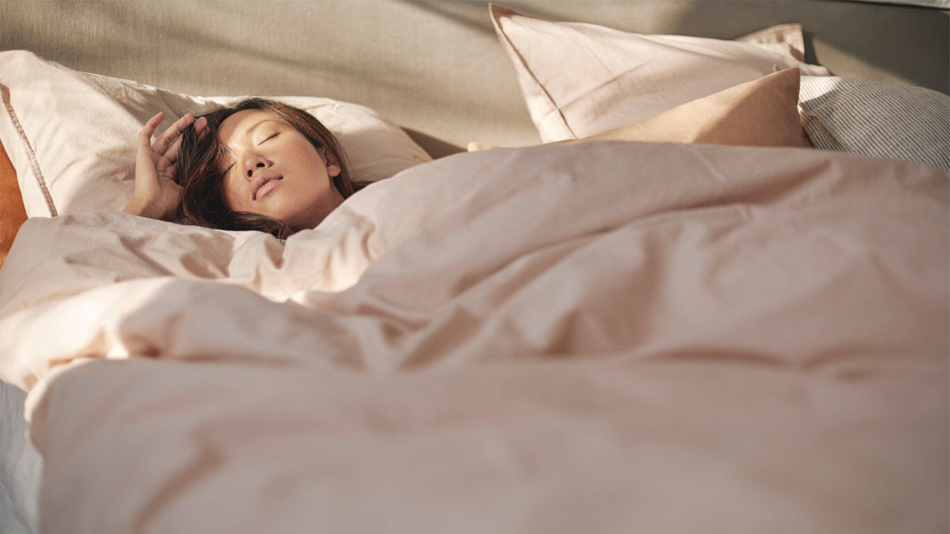 Frau liegt unter einer beigen Bettdecke in einem Bett und ruht auf mehreren Kissen.