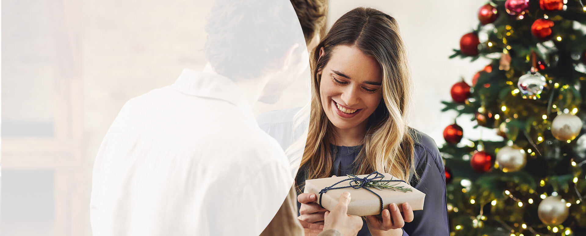 Mann übergibt Frau vor dem Weihnachtsbaum ein Geschenk.