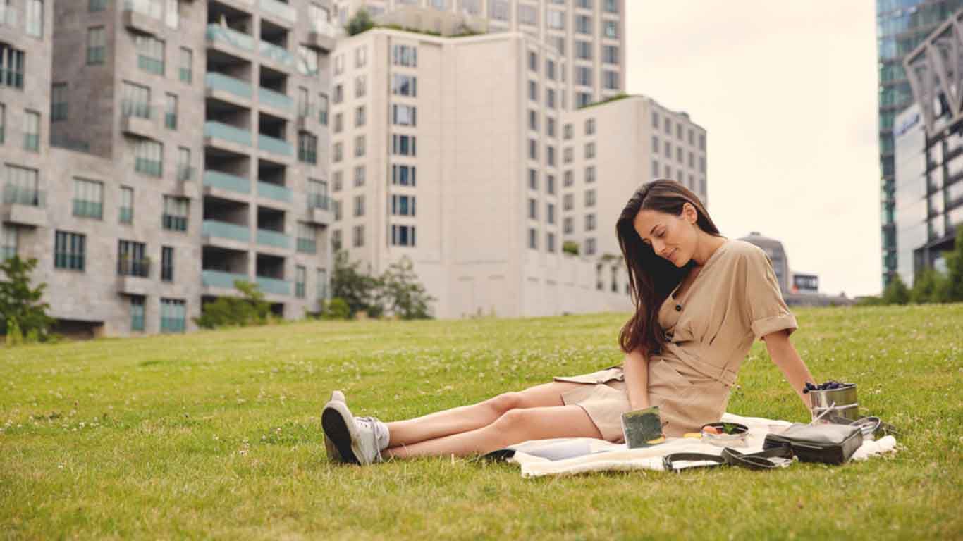 Frau in beigem Kleid sitzt mit Decke und B&uuml;chern auf einer Wiese. Im Hintergrund sind Hochh&auml;user zu sehen.