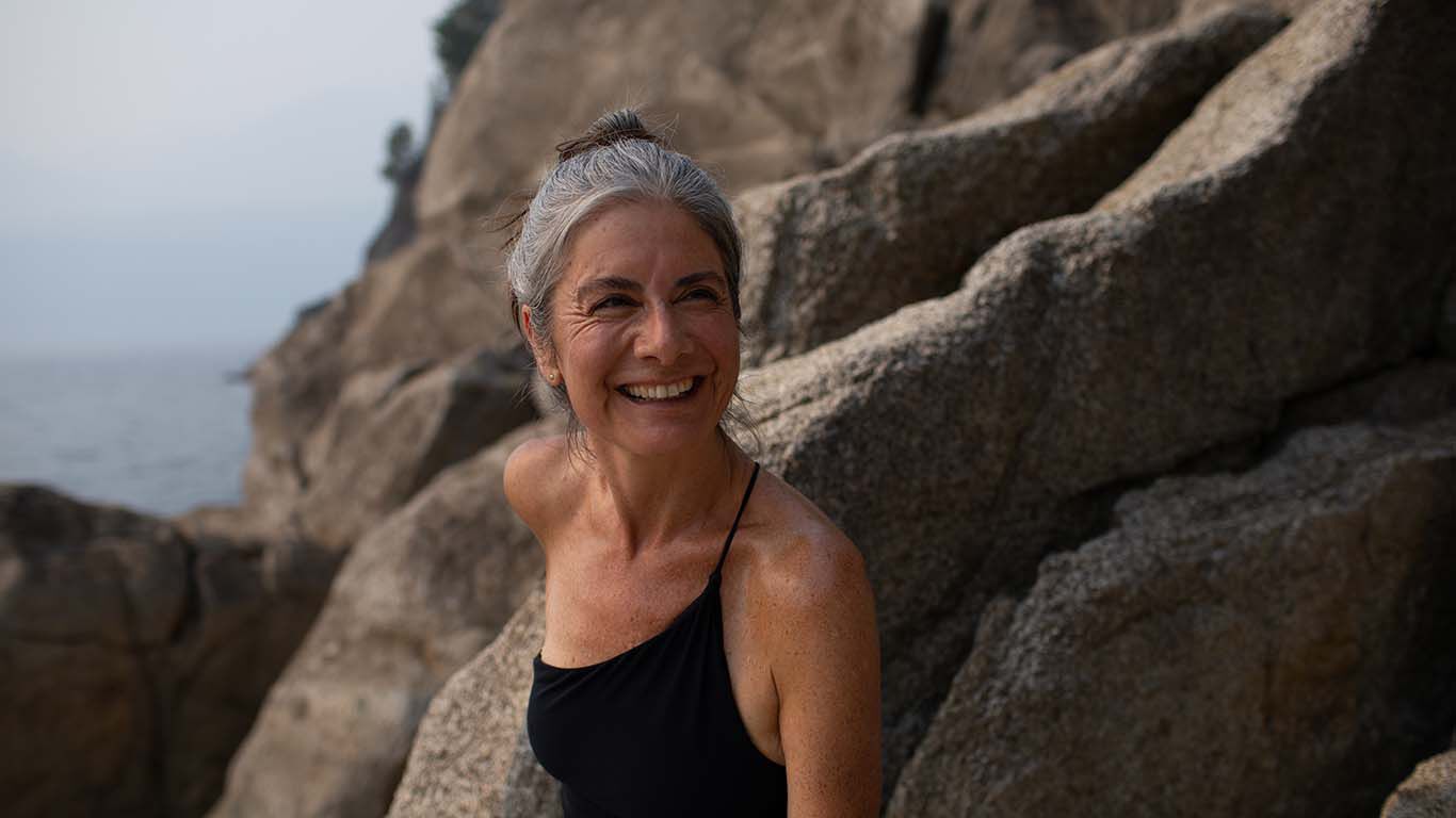 Dame steht im Badeanzug vor Felsen am Strand und lacht herzlich