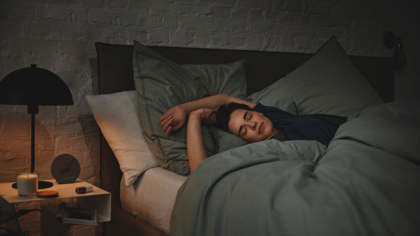 Frau liegt schlafend in einem Bett mit Kissen und Bettdecke in einem Schlafzimmer mit Nachttisch und Lampe.
