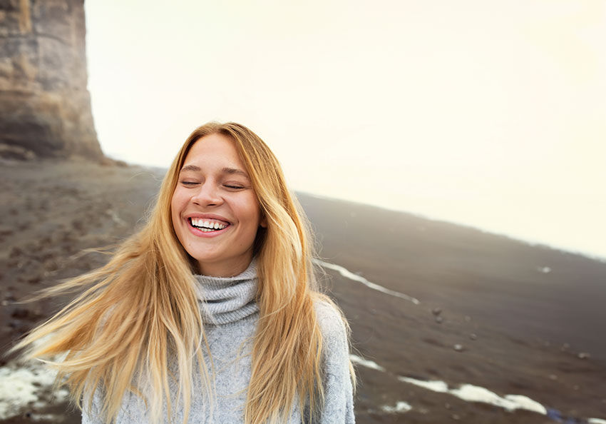 Blonde, junge Frau steht im Pullover am Strand und lacht. 