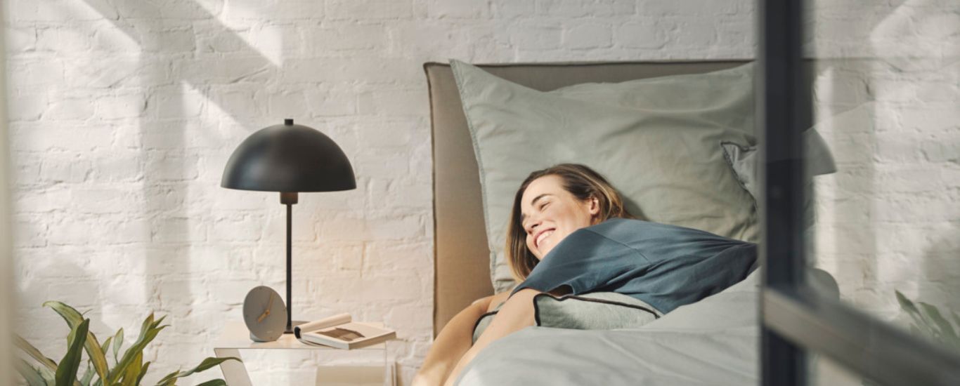  Frau liegt im Bett, l&auml;sst ihre Arme vom Bett h&auml;ngen und schaut in die Sonnenstrahlen.