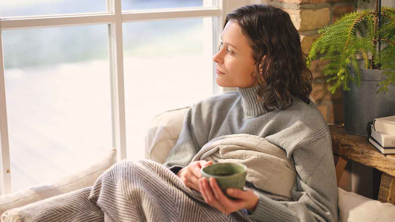 Frau sitzt mit Teetasse in Decke gewickelt am Fenster.