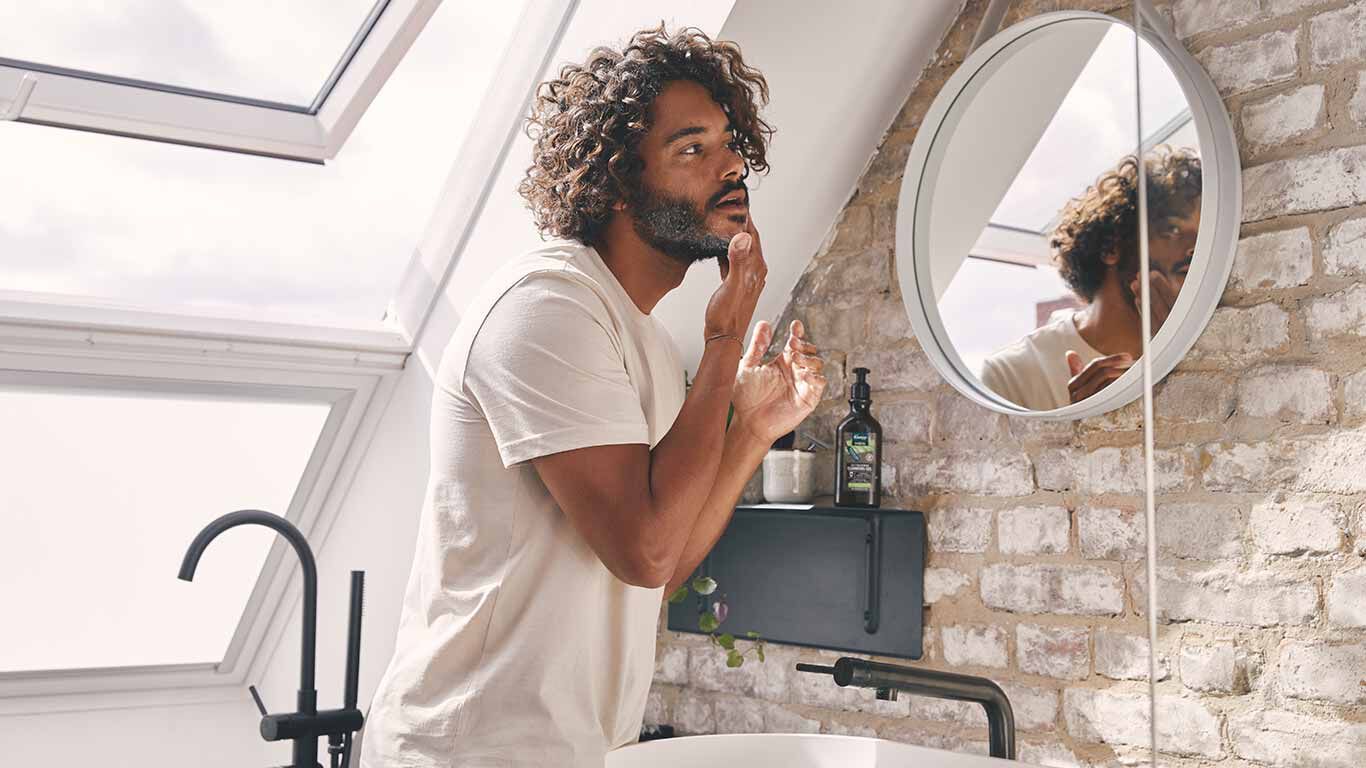 Mann mit dunklen Locken steht im Badezimmer vor dem Spiegel und pflegt seinen Bart.