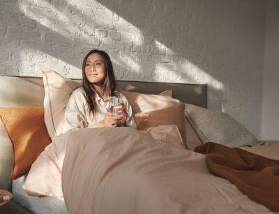  Frau sitzt mit einem Glas Wasser in den H&auml;nden aufrecht in einem Bett und die Sonne strahlt von au&szlig;en auf sie herein.