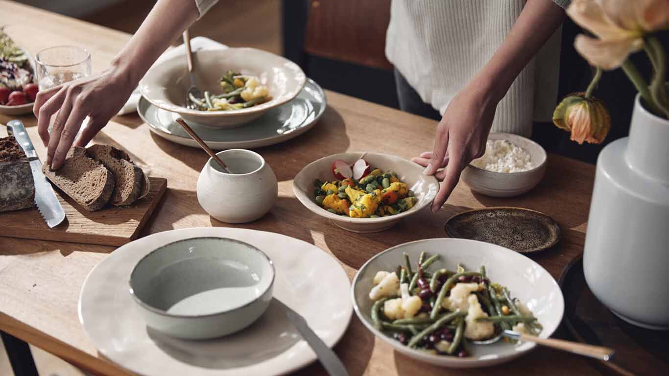 Gedeckter Tisch mit Sch&uuml;sseln, Brot und frischem Gem&uuml;se, H&auml;nde beim Servieren.&ldquo;
