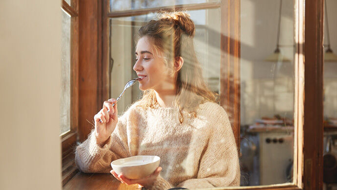 Frau sitzt mit gesunden Fr&uuml;hst&uuml;ck am Fenster mit einer Sch&uuml;ssel M&uuml;sli. Nat&uuml;rliches Tageslicht und eine entspannte Atmosph&auml;re.