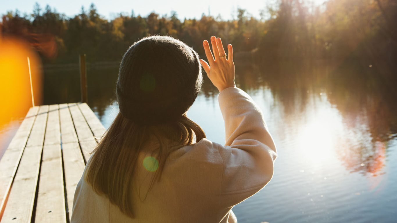 Frau steht an einem Steg und hält sich mit der Hand die Sonne aus dem Gesicht. 